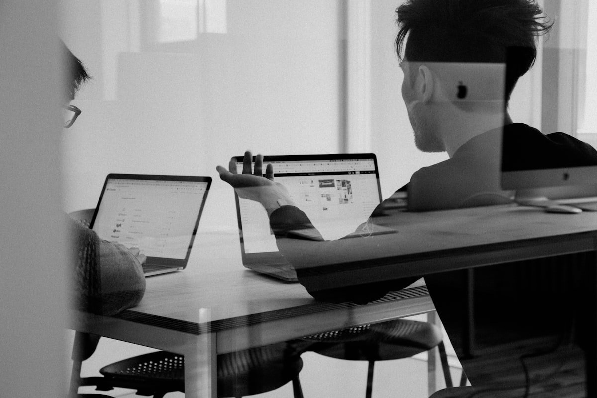 Duas pessoas sentadas em uma mesa trabalhando em laptops, uma delas gesticulando em direção à tela, em um ambiente de escritório moderno.