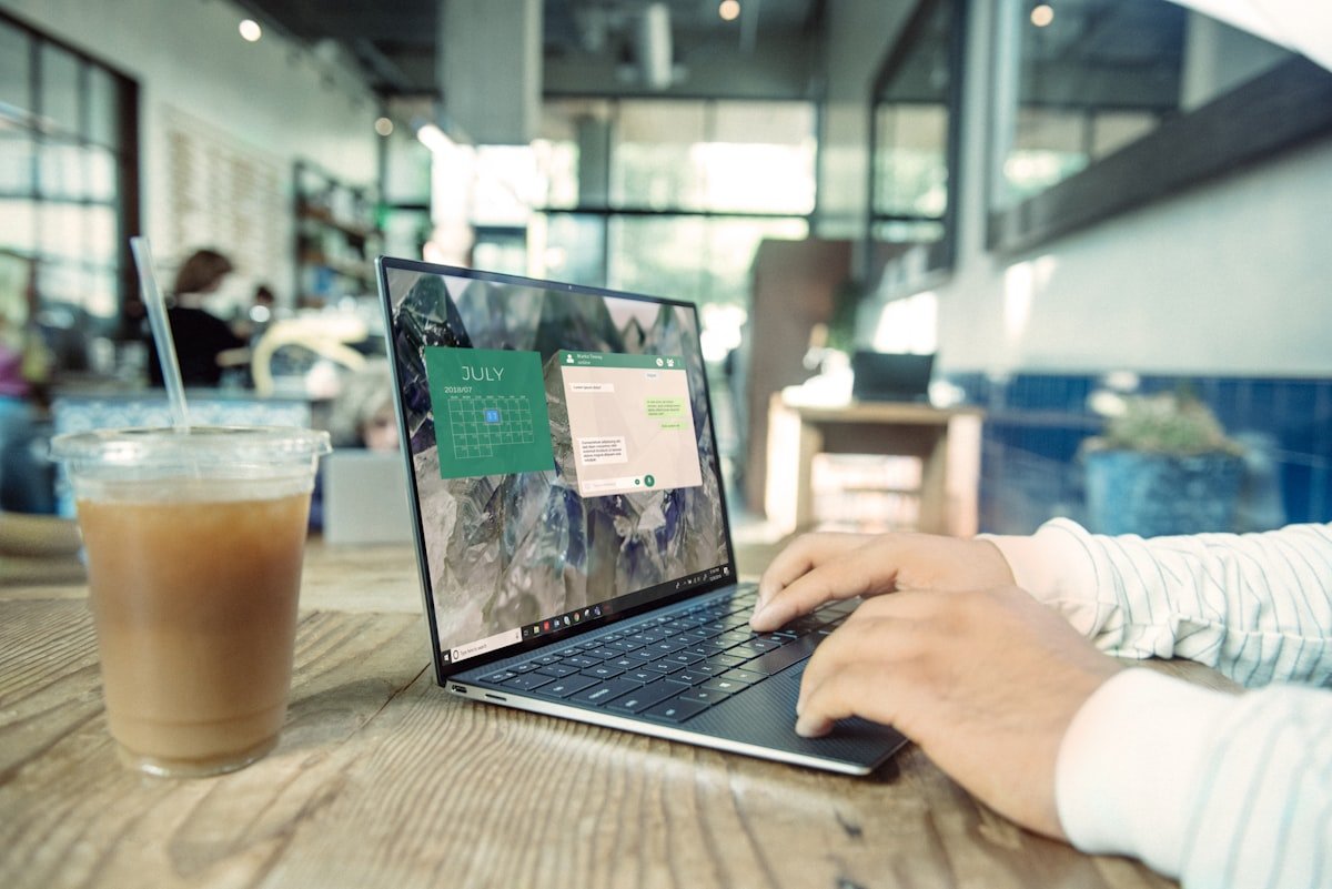 Pessoa digitando em um laptop em uma mesa de madeira em um café, com um café gelado por perto e um aplicativo de calendário aberto na tela do laptop.