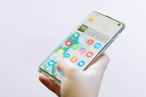 Uma mão segurando um smartphone com vários ícones de aplicativos visíveis na tela, incluindo aplicativos de mídia social e de mensagens.