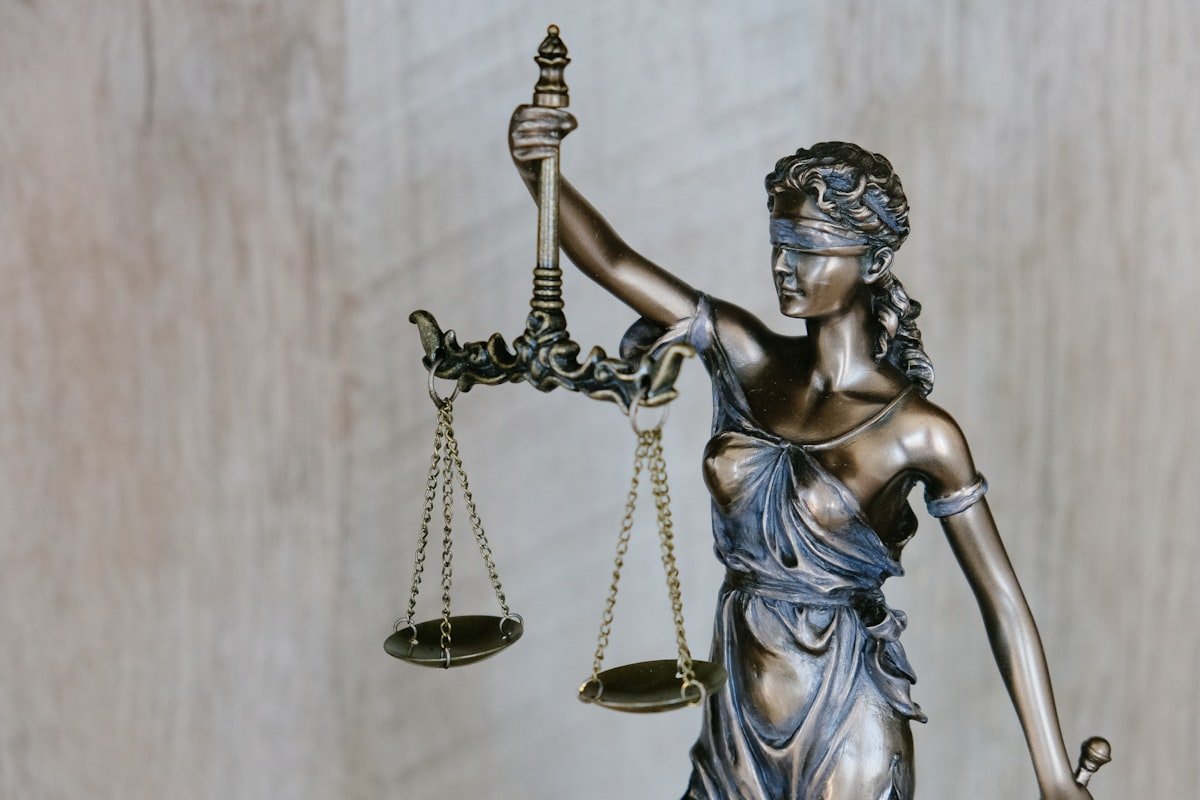 Estátua de bronze da Lady Justice segurando uma balança equilibrada na mão esquerda e uma espada na direita, usando uma venda nos olhos em um fundo neutro.