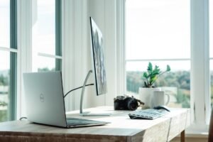 Uma mesa com um laptop Dell, um monitor externo, um teclado sem fio, uma câmera, uma caneca de café e um vaso de plantas perto de grandes janelas com vista panorâmica.