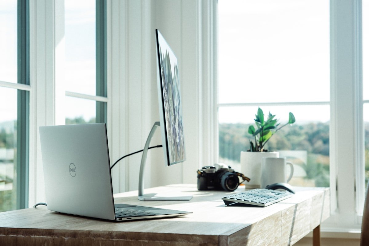 Uma mesa com um laptop Dell, um monitor externo, um teclado sem fio, uma câmera, uma caneca de café e um vaso de plantas perto de grandes janelas com vista panorâmica.