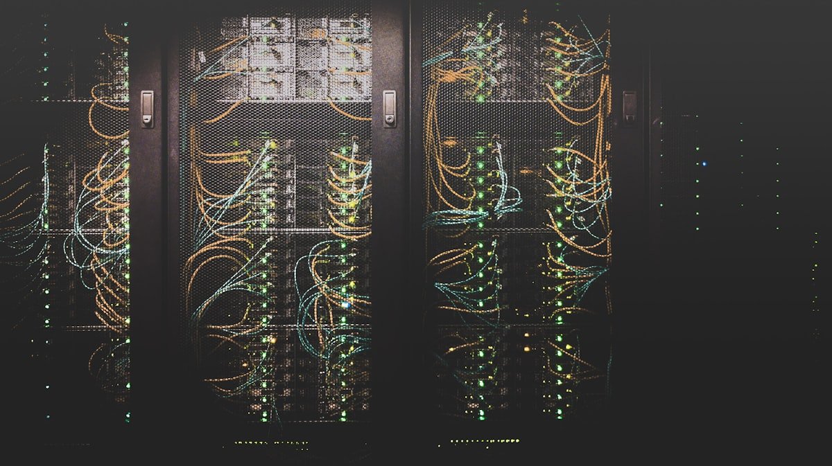 Fileiras de servidores de rede com cabos visíveis e luzes indicadoras estão alojadas em um rack escuro de data center.