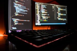 Um teclado de computador em primeiro plano com dois monitores exibindo linhas coloridas de código em uma sala escura.