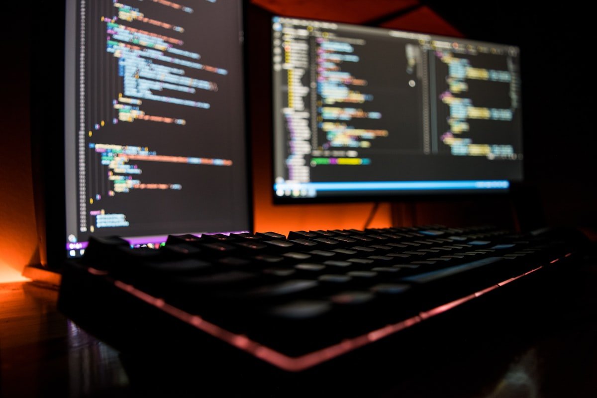 Um teclado de computador em primeiro plano com dois monitores exibindo linhas coloridas de código em uma sala escura.