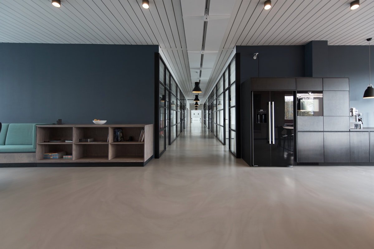Interior de escritório moderno com piso polido, paredes escuras, uma área de estar à esquerda, escritórios com paredes de vidro ao longo de um corredor central e uma cozinha compacta à direita.